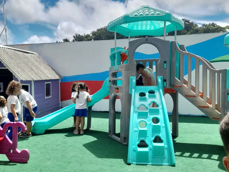O pátio da Escola Magia num dia de sol, com o brinquedo grande, o escorregador e a casinha lilás, e crianças brincando ao fundo