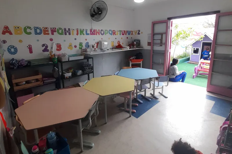 Uma sala da Escola Magia pela manhã, com as mesas coloridas, o alfabeto na parede e a porta aberta para o pátio