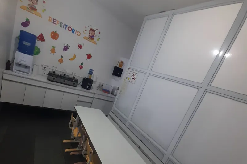 O refeitório da Escola Magia, com a mesa comprida, as cadeiras pequenas e frutas desenhadas na parede