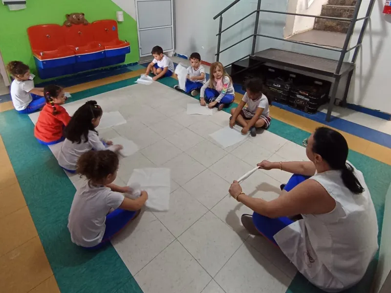 Crianças sentadas em roda no chão da sala, cada uma com uma folha, fazendo a atividade com a professora